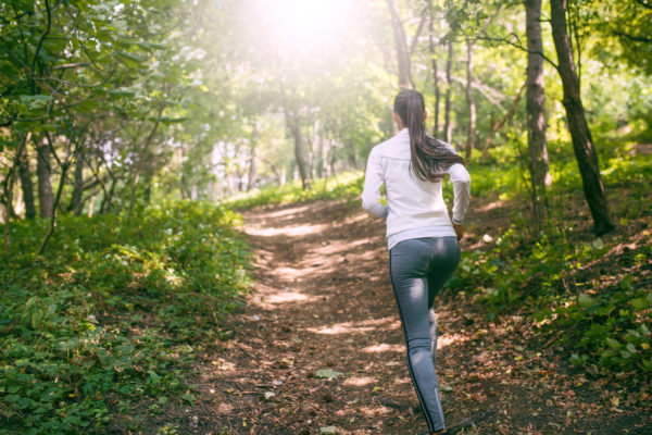 Beautiful view of Caucasian woman runner wearing grey leggings doing jogging exercises outdoors in forest. Action shot of female jogger with athletic body during morning run. Healthy motivation. 